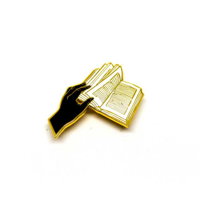 hard enamel pin, a hand holds open the pages of a book, the words in the book are represented by abstract, straight lines, gold outlines on black hand, white book