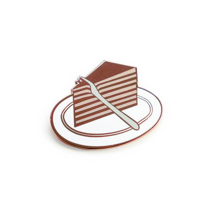 a hard enamel pin, grey fork cutting through a wedged slice of pink and copper layered cake on a white plate